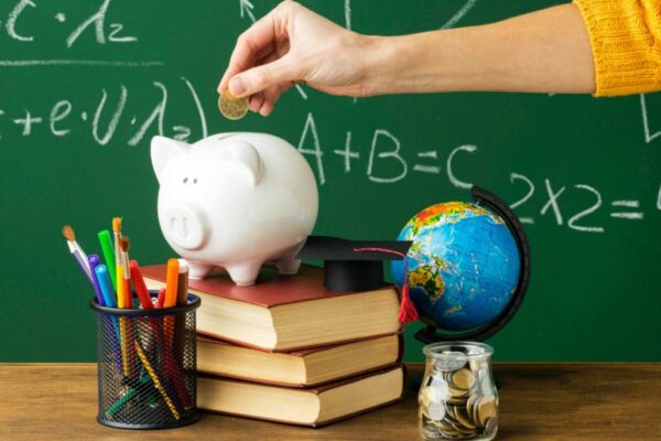 Uma mão coloca uma moeda em um cofrinho de porquinho, em frente a um quadro verde com equações matemáticas e um globo terrestre. Há livros e um porta-canetas com canetas coloridas na mesa.