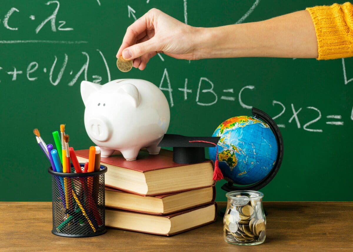 Uma mão coloca uma moeda em um cofrinho de porquinho, em frente a um quadro verde com equações matemáticas e um globo terrestre. Há livros e um porta-canetas com canetas coloridas na mesa.