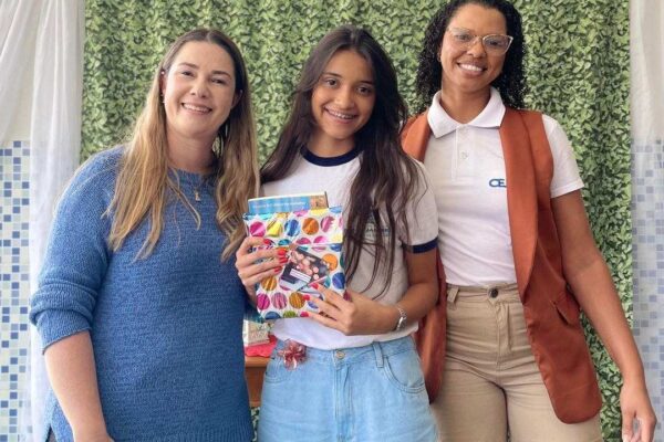 Aluna recebe livro com sua redação publicada, acompanhada por professoras, em evento da Seeduc-RJ.