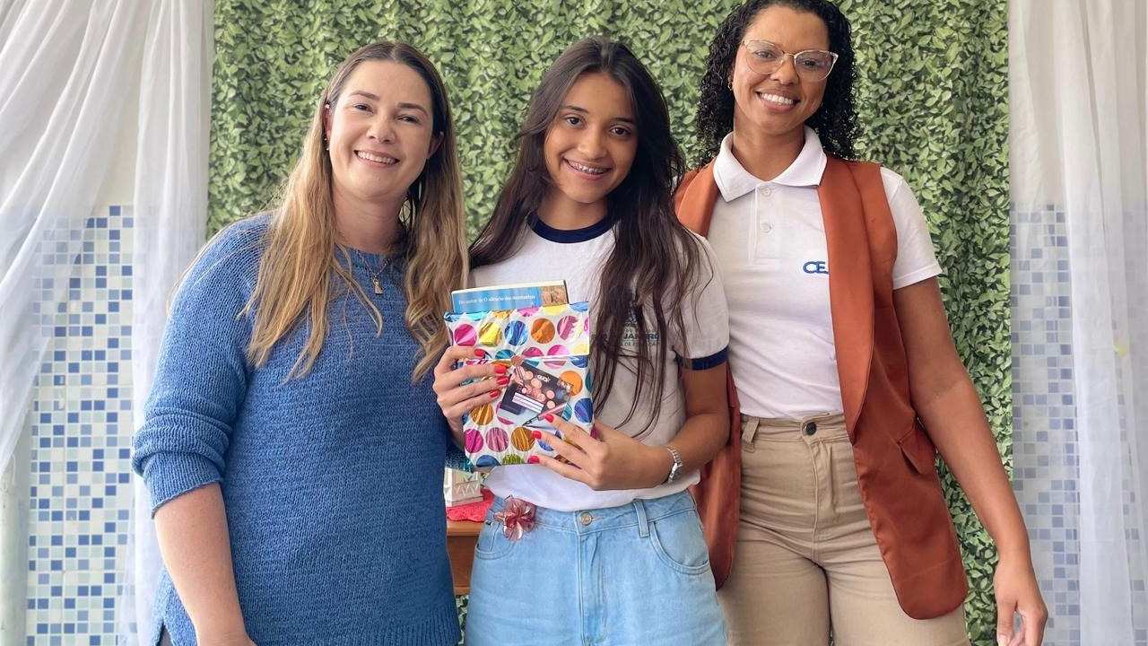 Aluna recebe livro com sua redação publicada, acompanhada por professoras, em evento da Seeduc-RJ.