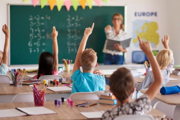 Alunos levantando a mão em sala de aula, com professora e lousa com contas.