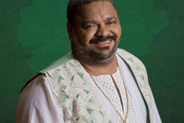 Arlindo Cruz sorrindo, com roupas claras e corrente dourada, fundo verde