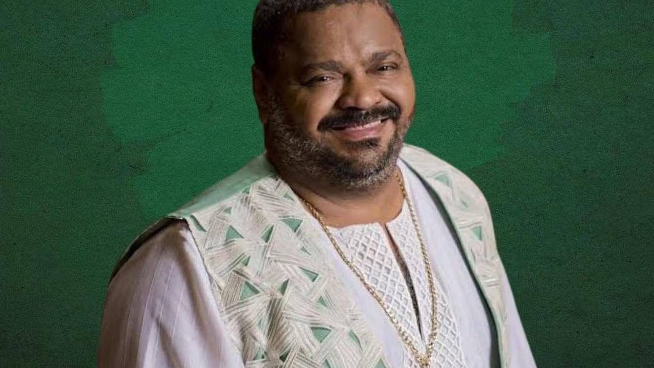 Arlindo Cruz sorrindo, com roupas claras e corrente dourada, fundo verde