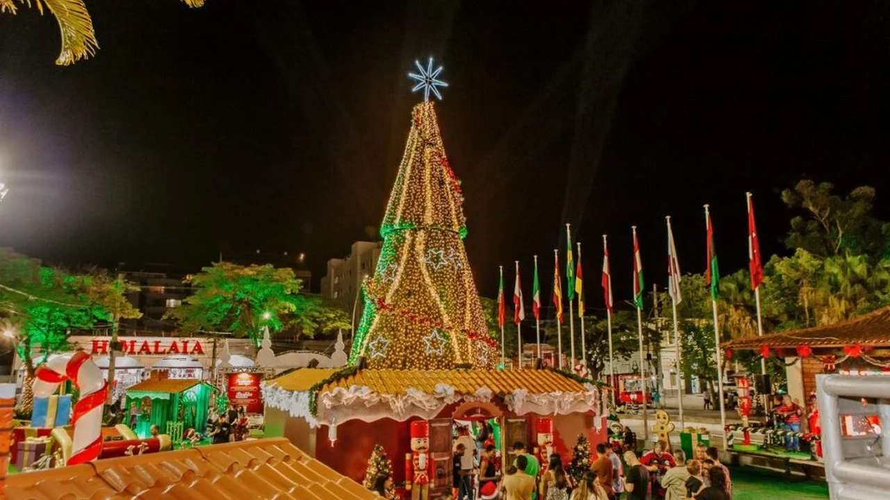 Árvore de Natal iluminada em praça pública, com enfeites e pessoas ao redor.