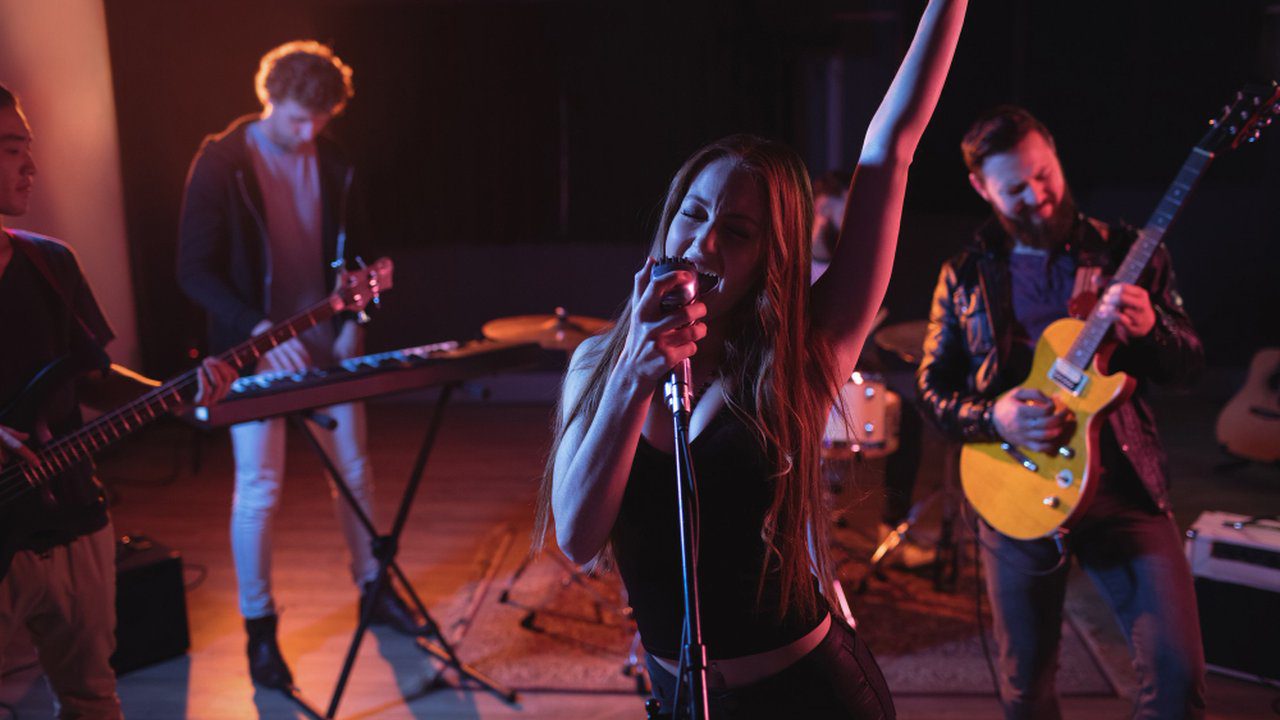 Banda de rock ao vivo com vocalista feminina cantando no palco, com outros músicos tocando instrumentos.