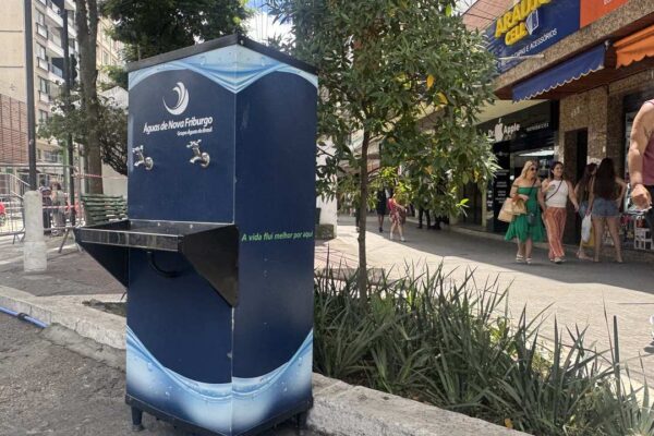 Bebedouro da Águas de Nova Friburgo em área urbana, com pessoas ao fundo e árvores.
