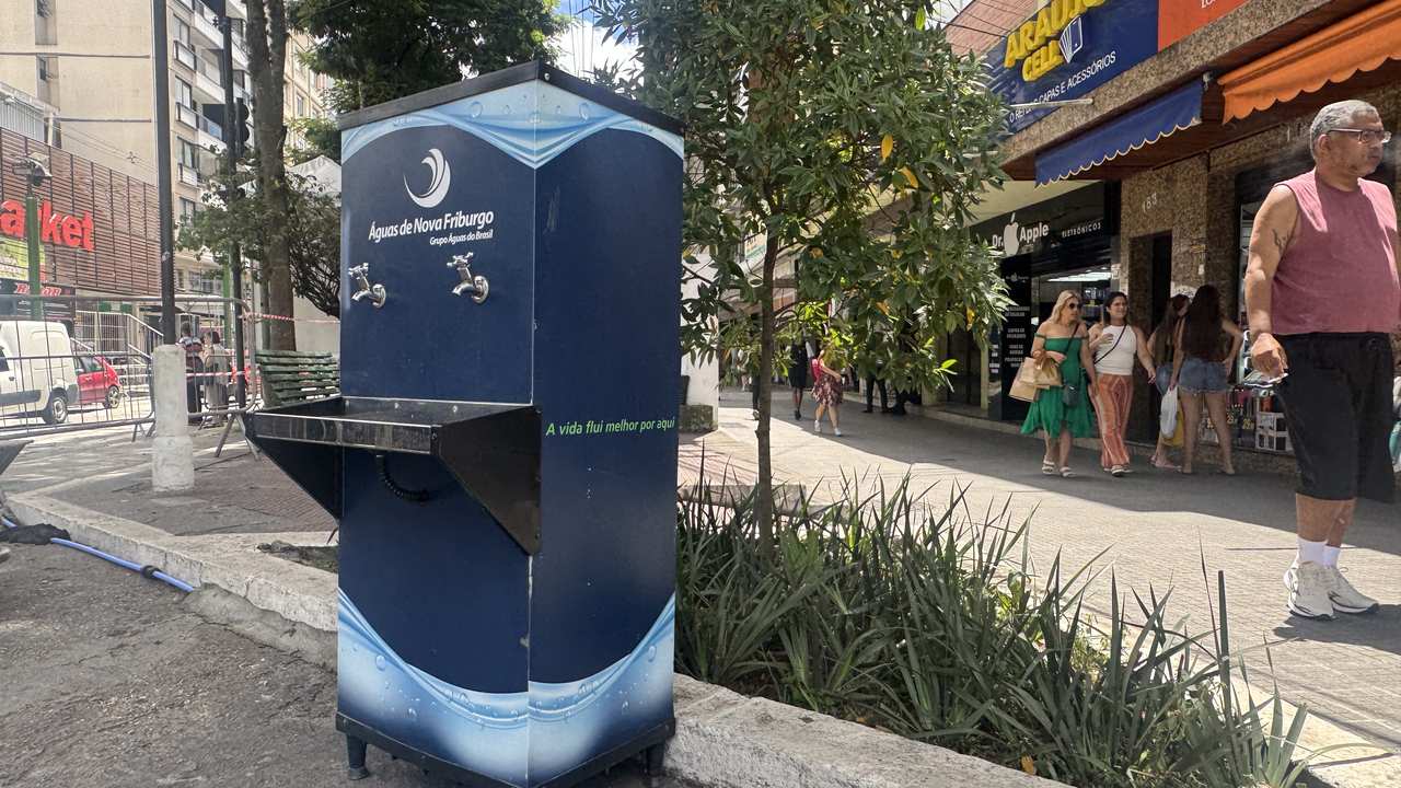 Bebedouro da Águas de Nova Friburgo em área urbana, com pessoas ao fundo e árvores.
