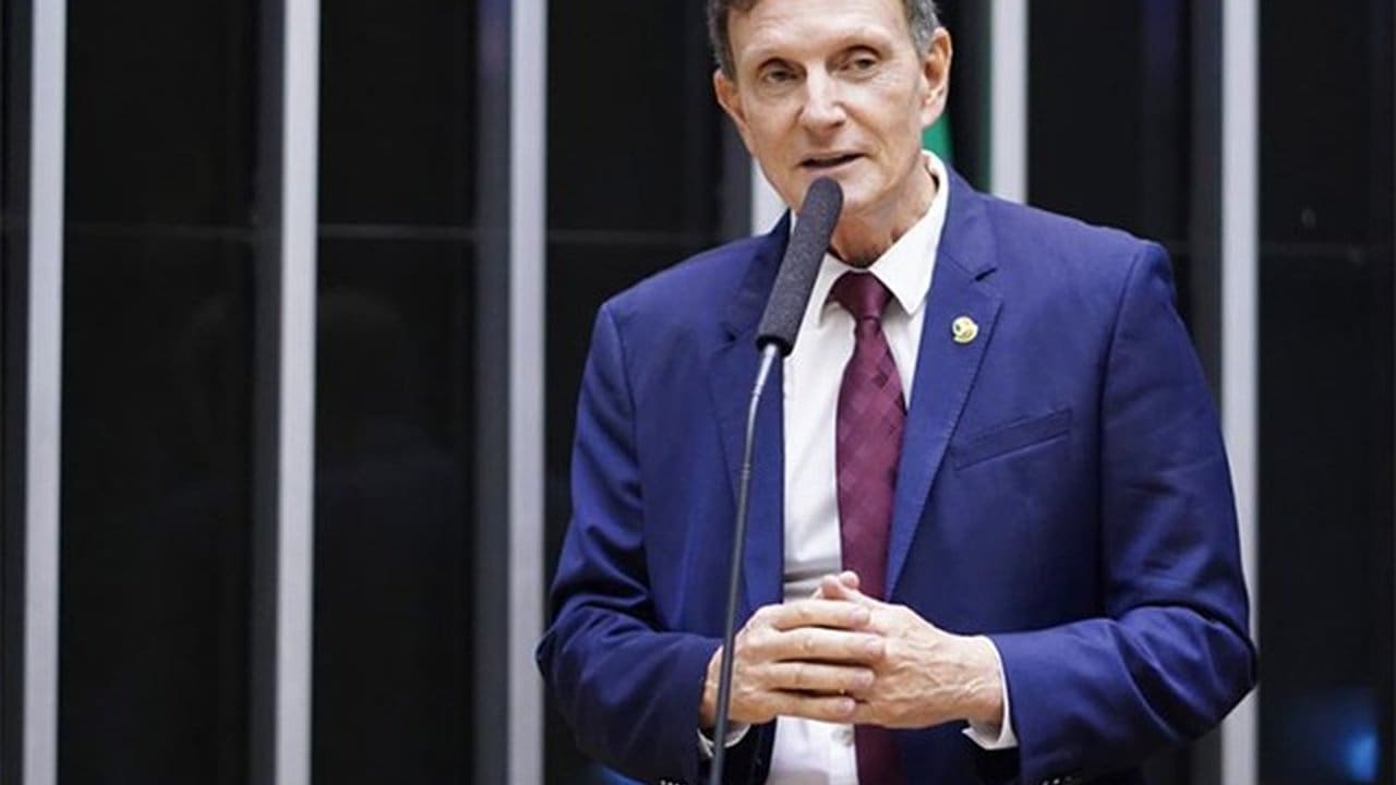 Bispo Marcelo Crivella falando em um evento, com um microfone na frente e um terno azul.