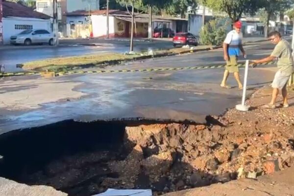 Buraco enorme em rua após rompimento de tubulação, com pessoas observando.