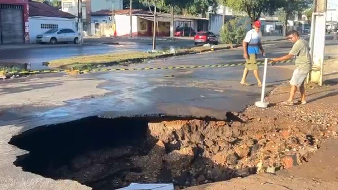 Buraco enorme em rua após rompimento de tubulação, com pessoas observando.
