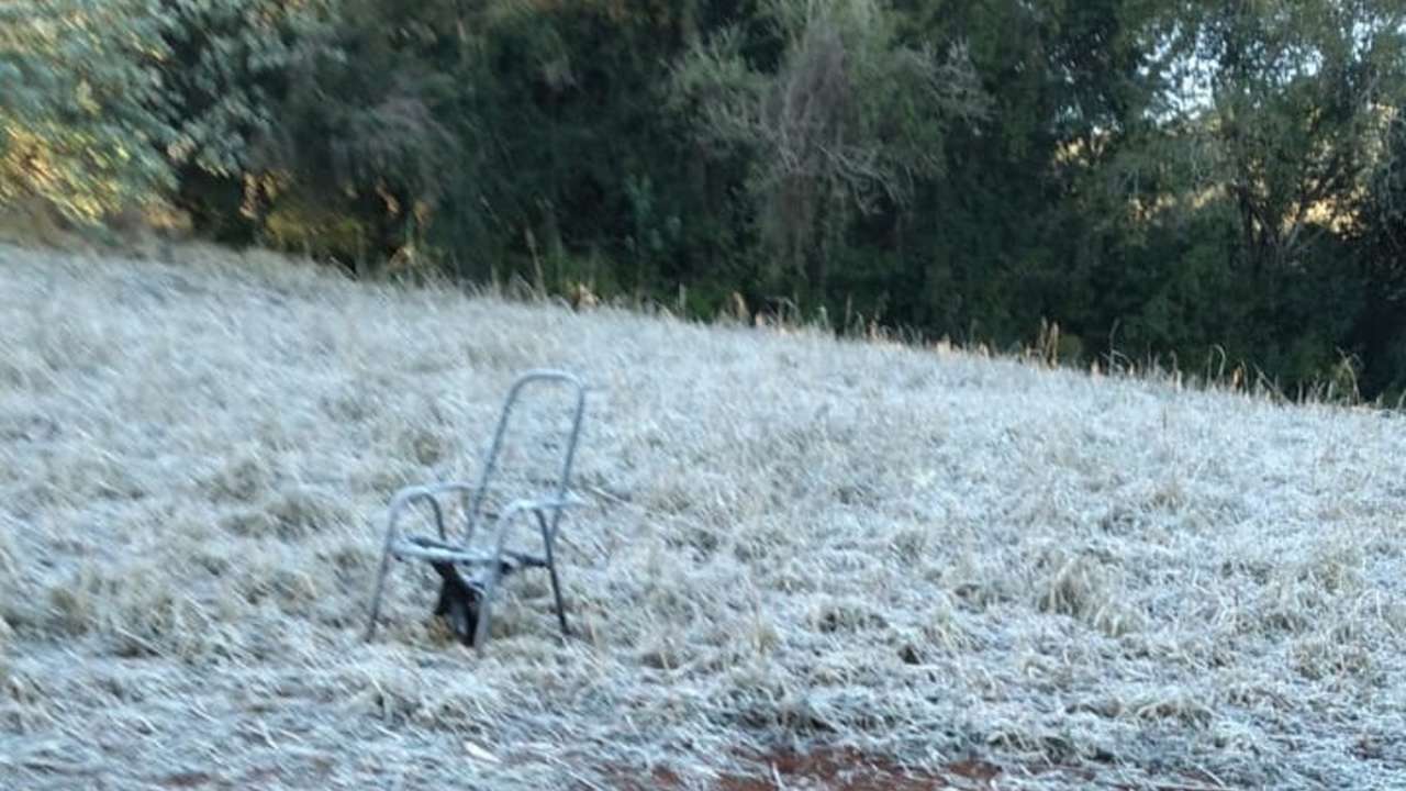 Cadeira de metal enferrujada em campo coberto por geada, com árvores ao fundo.