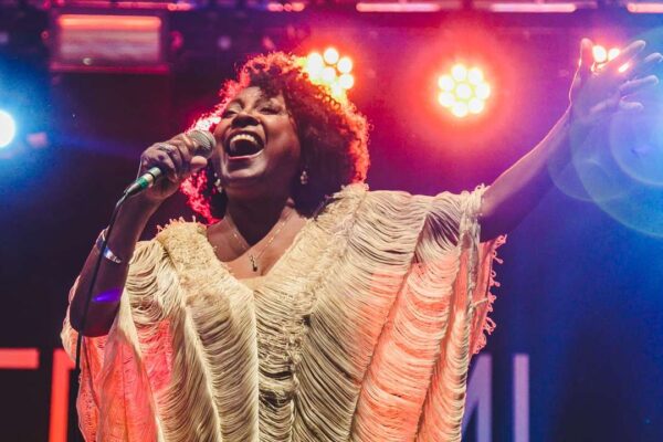 Cantora de cabelos crespos em um show, usando um vestido branco com detalhes em franjas, cantando em um microfone e olhando para a frente.