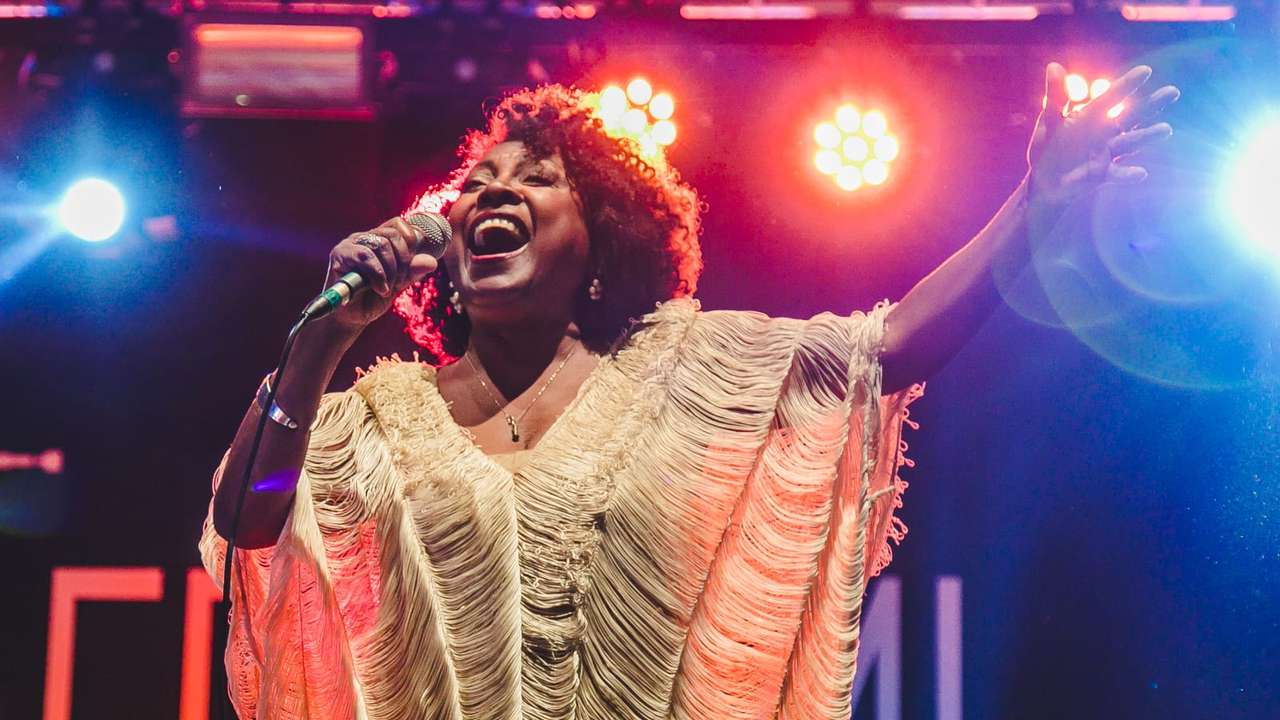 Cantora de cabelos crespos em um show, usando um vestido branco com detalhes em franjas, cantando em um microfone e olhando para a frente.