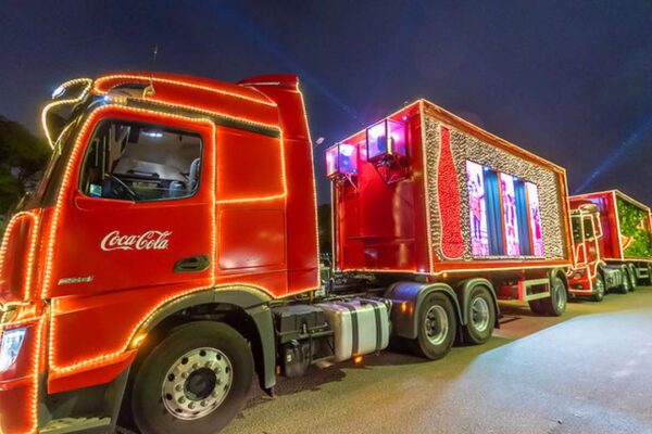 Caravana de Natal da Coca-Cola com caminhões decorados com luzes e telas exibindo imagens natalinas.