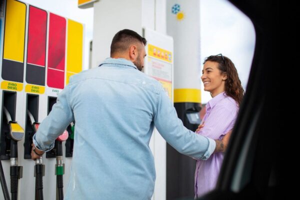 Casal abastecendo carro em posto de gasolina, com homem segurando a mangueira e mulher sorrindo.