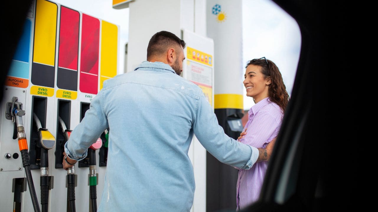 Casal abastecendo carro em posto de gasolina, com homem segurando a mangueira e mulher sorrindo.