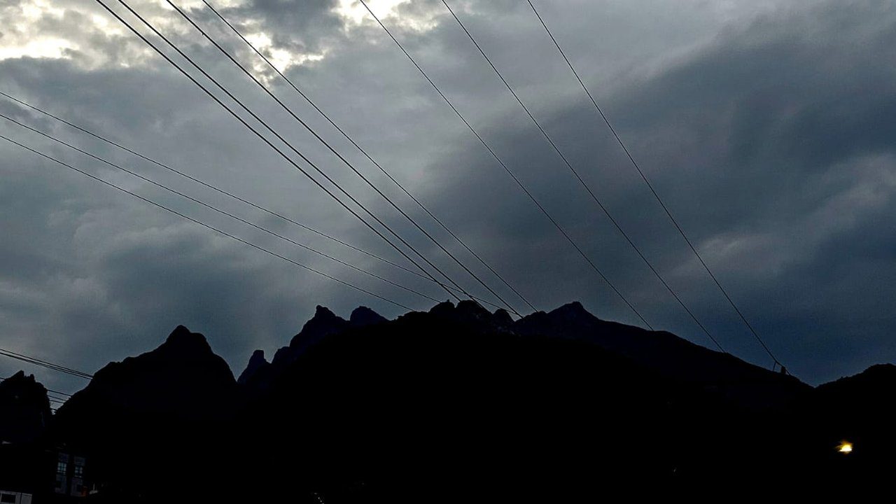 Céu nublado com silhueta de montanhas e cabos de energia, sob efeito da frente fria.