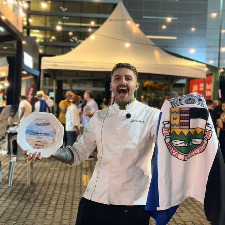 Chef João Vieira, de Nova Friburgo, sorri segurando um prêmio e uma bandeira, comemorando o título de Melhor Chef do Brasil em 2025.
