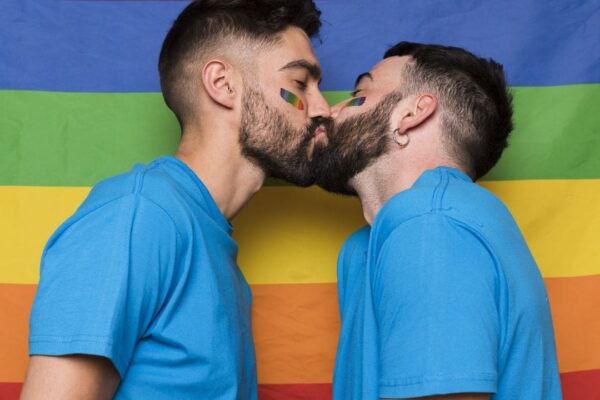 Dois homens se beijando em frente à bandeira do arco-íris, simbolizando orgulho LGBTQIA+.