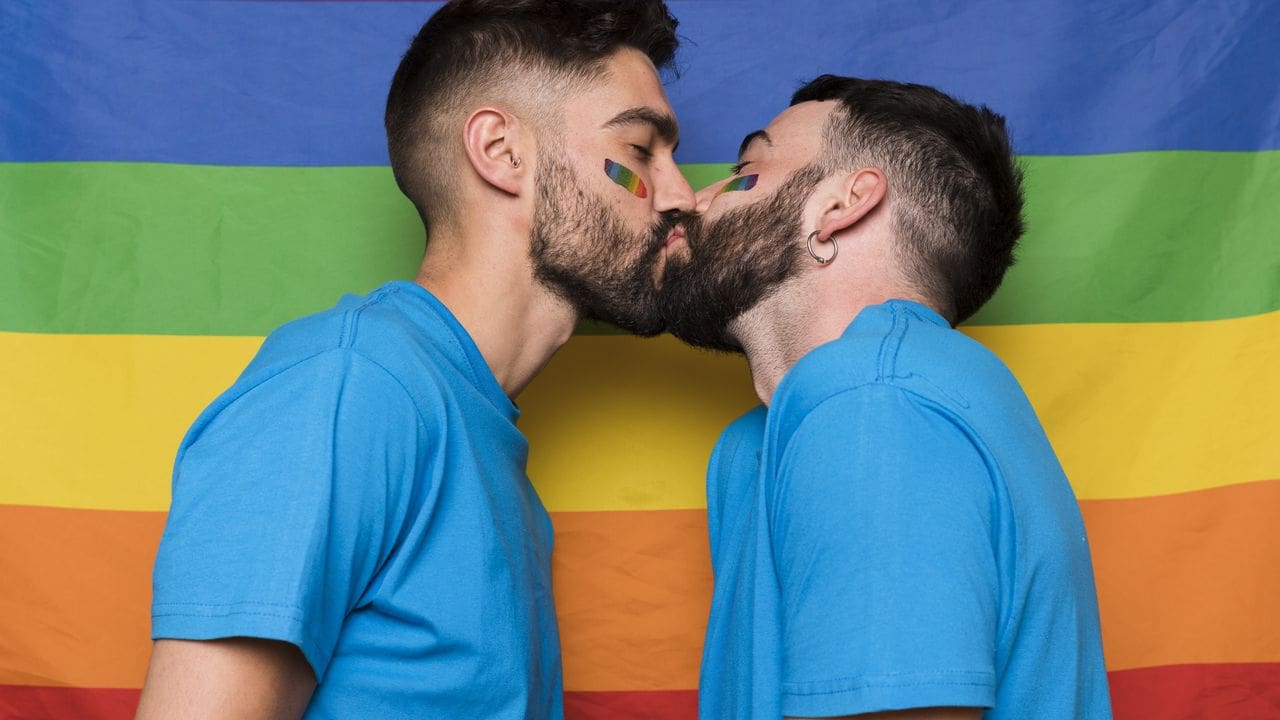 Dois homens se beijando em frente à bandeira do arco-íris, simbolizando orgulho LGBTQIA+.