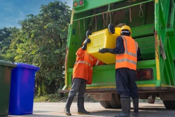 Dois trabalhadores de coleta de lixo descarregando um recipiente amarelo em um caminhão de lixo verde.