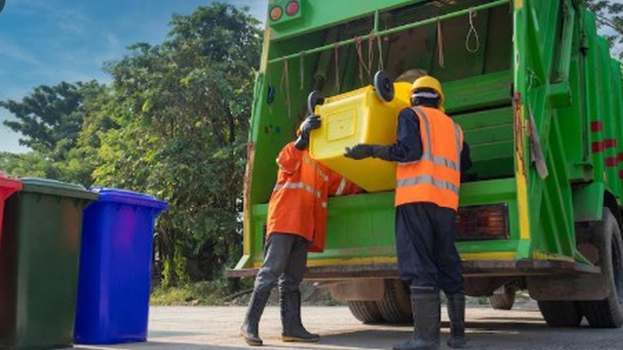 Dois trabalhadores de coleta de lixo descarregando um recipiente amarelo em um caminhão de lixo verde.