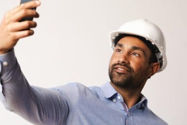 Engenheiro civil com capacete branco tirando selfie com celular.