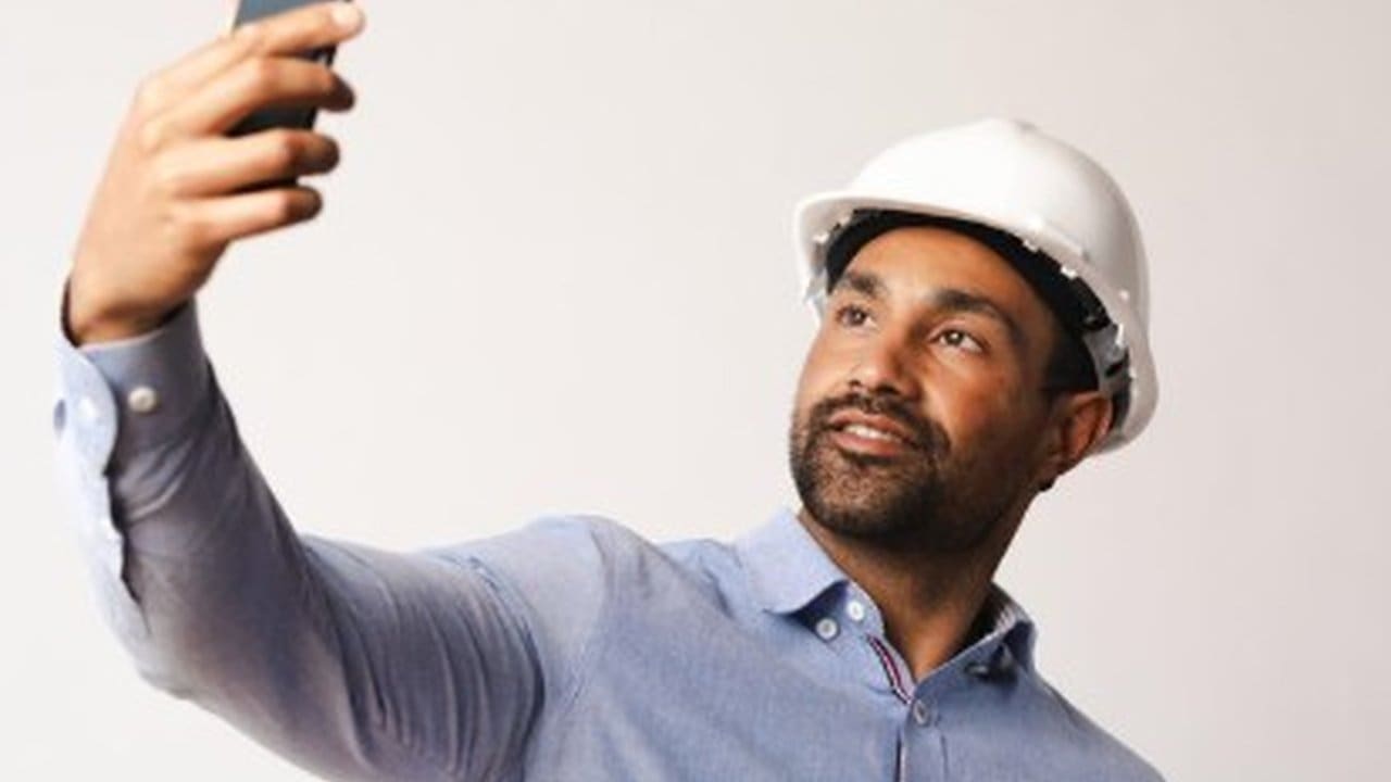Engenheiro civil com capacete branco tirando selfie com celular.