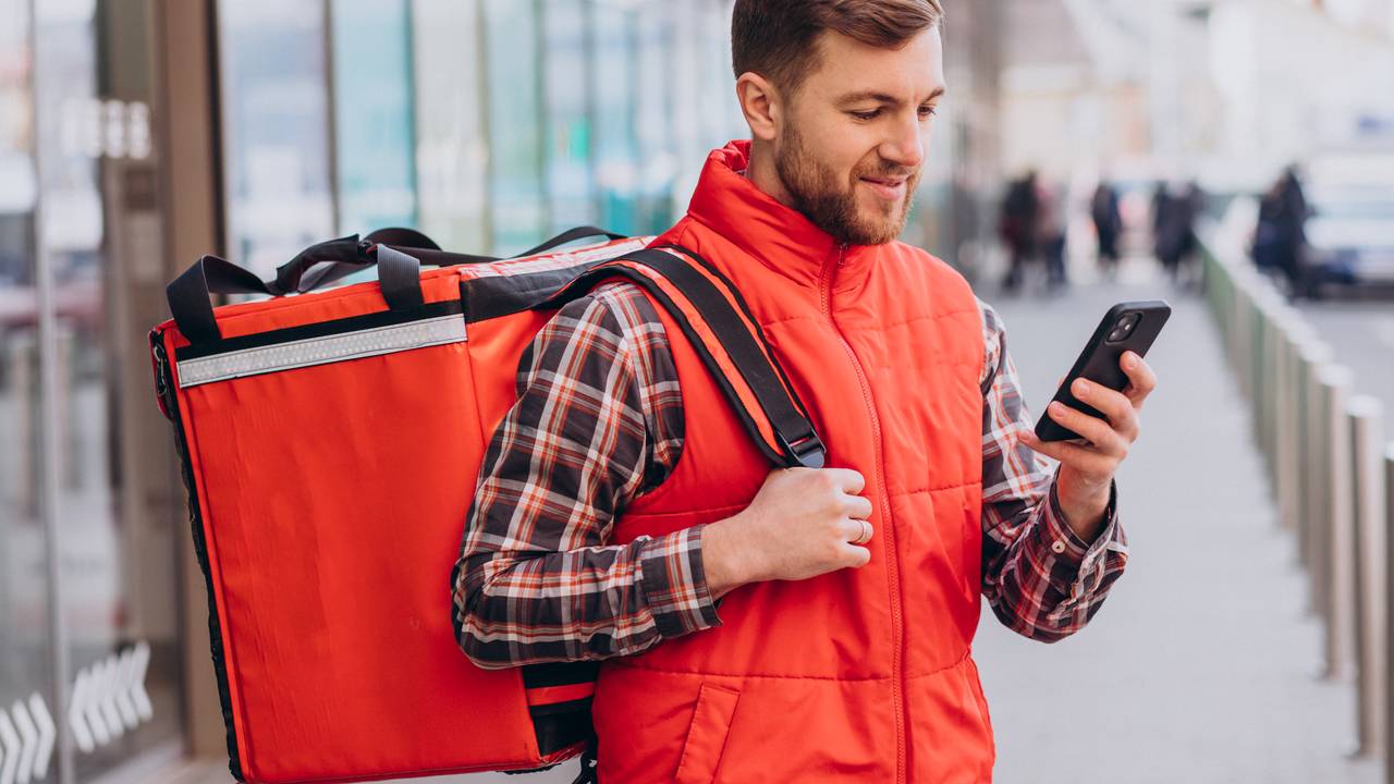 Entregador com mochila térmica e celular em frente a um prédio, no Rio de Janeiro.