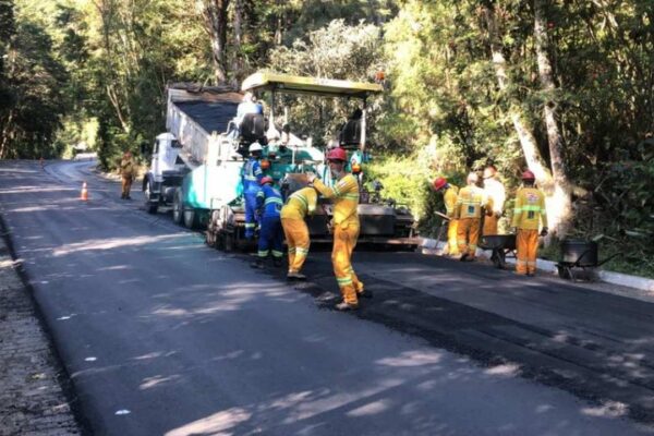 Equipe de obras trabalhando na pavimentação de estrada, com máquina e árvores ao fundo.