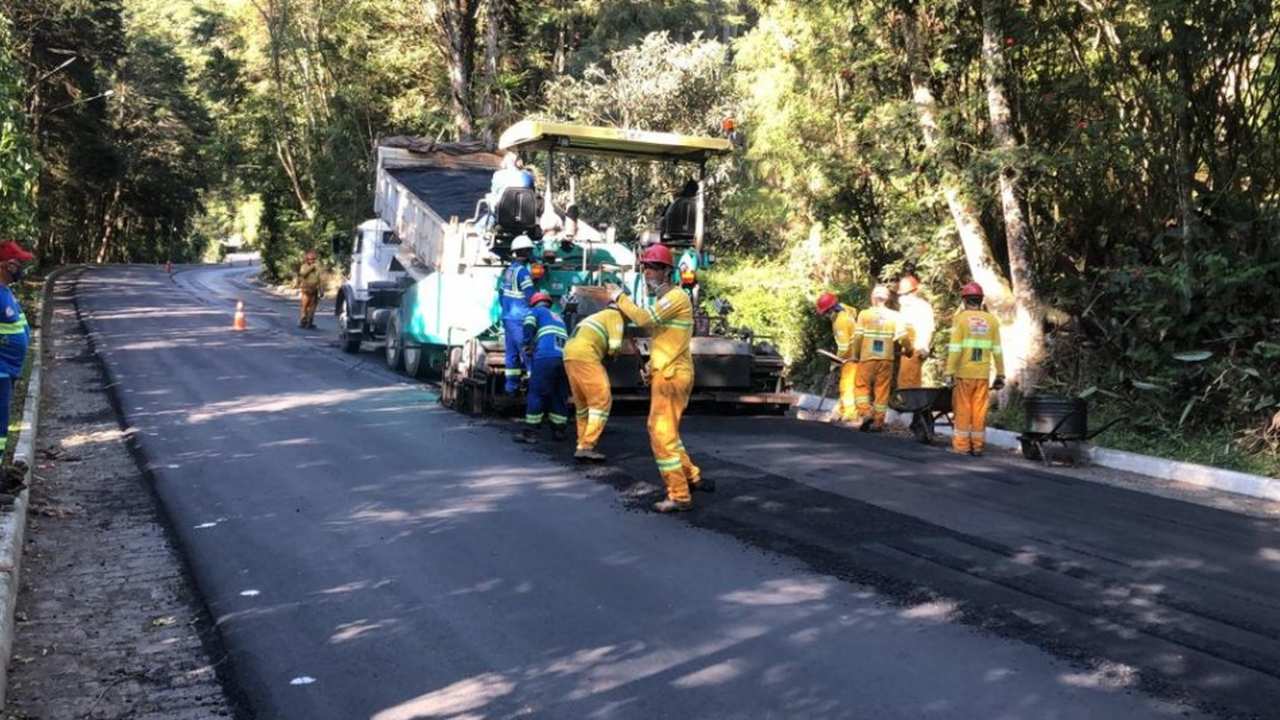 Equipe de obras trabalhando na pavimentação de estrada, com máquina e árvores ao fundo.