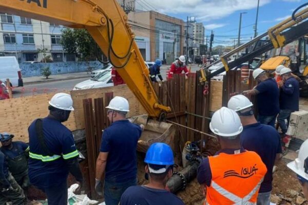 Equipe trabalhando em reparo de tubulação de água, com máquina e vários trabalhadores uniformizados.