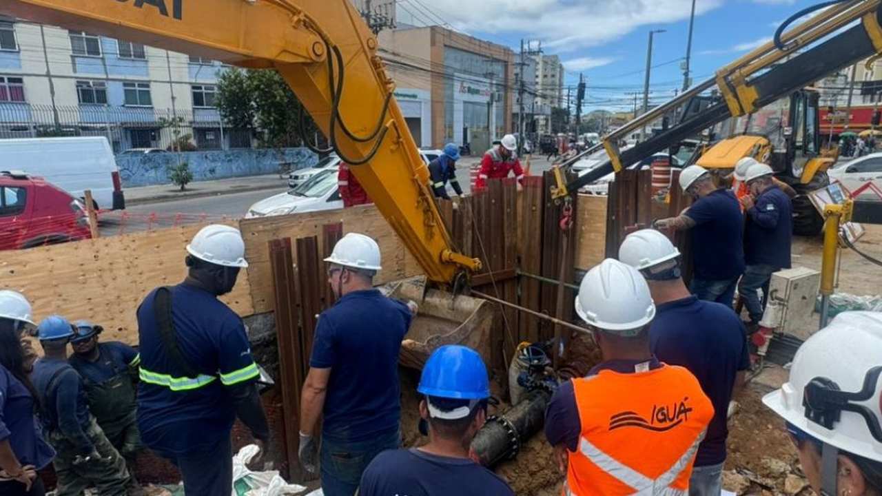 Equipe trabalhando em reparo de tubulação de água, com máquina e vários trabalhadores uniformizados.