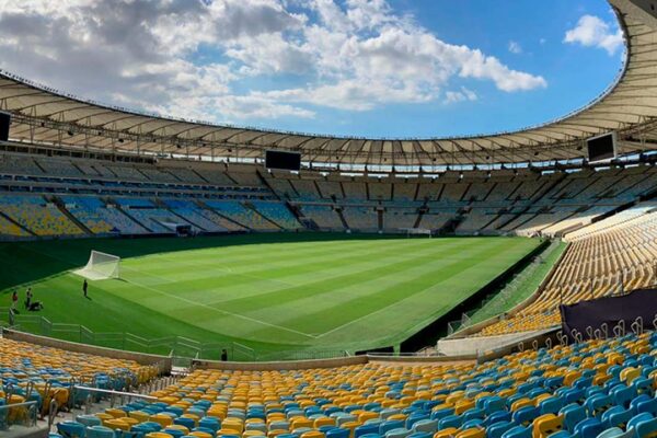 Estádio do Maracanã com gramado e arquibancadas vazias sob céu azul.