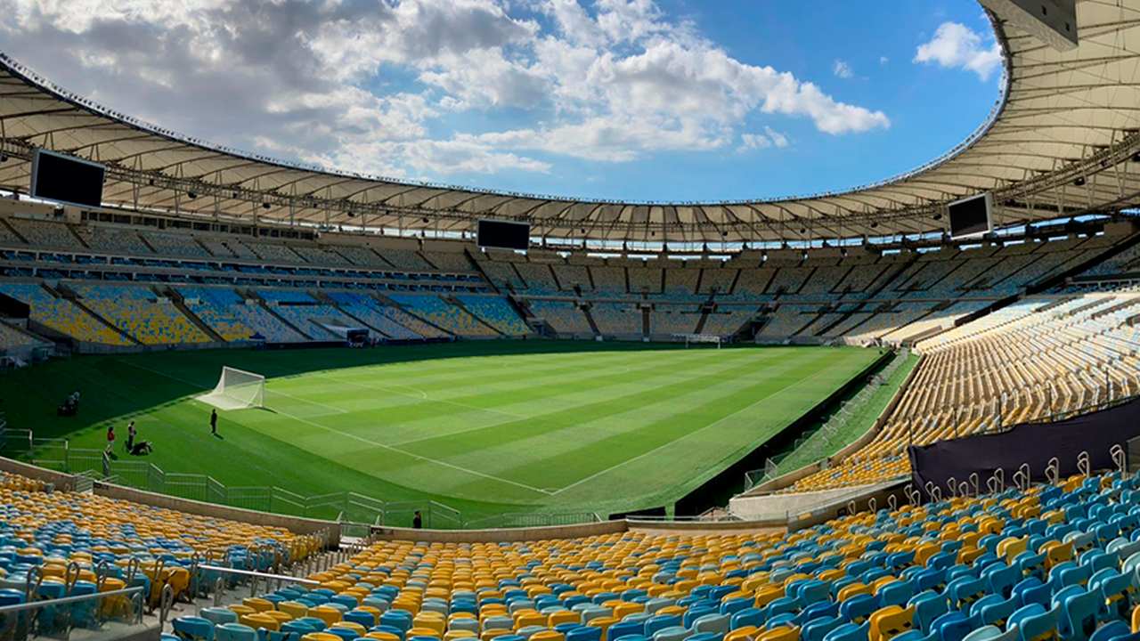 Estádio do Maracanã com gramado e arquibancadas vazias sob céu azul.