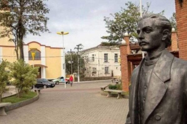 Estátua de Casimiro de Abreu em praça, com igreja ao fundo e cenário urbano.