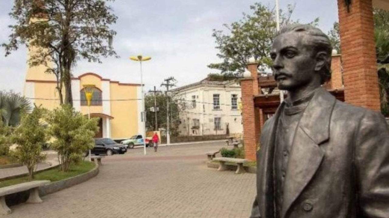 Estátua de Casimiro de Abreu em praça, com igreja ao fundo e cenário urbano.