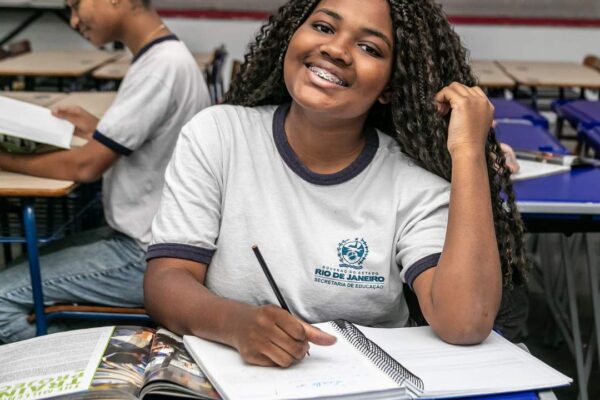 Estudante sorrindo com aparelho nos dentes, em sala de aula, escrevendo em caderno.