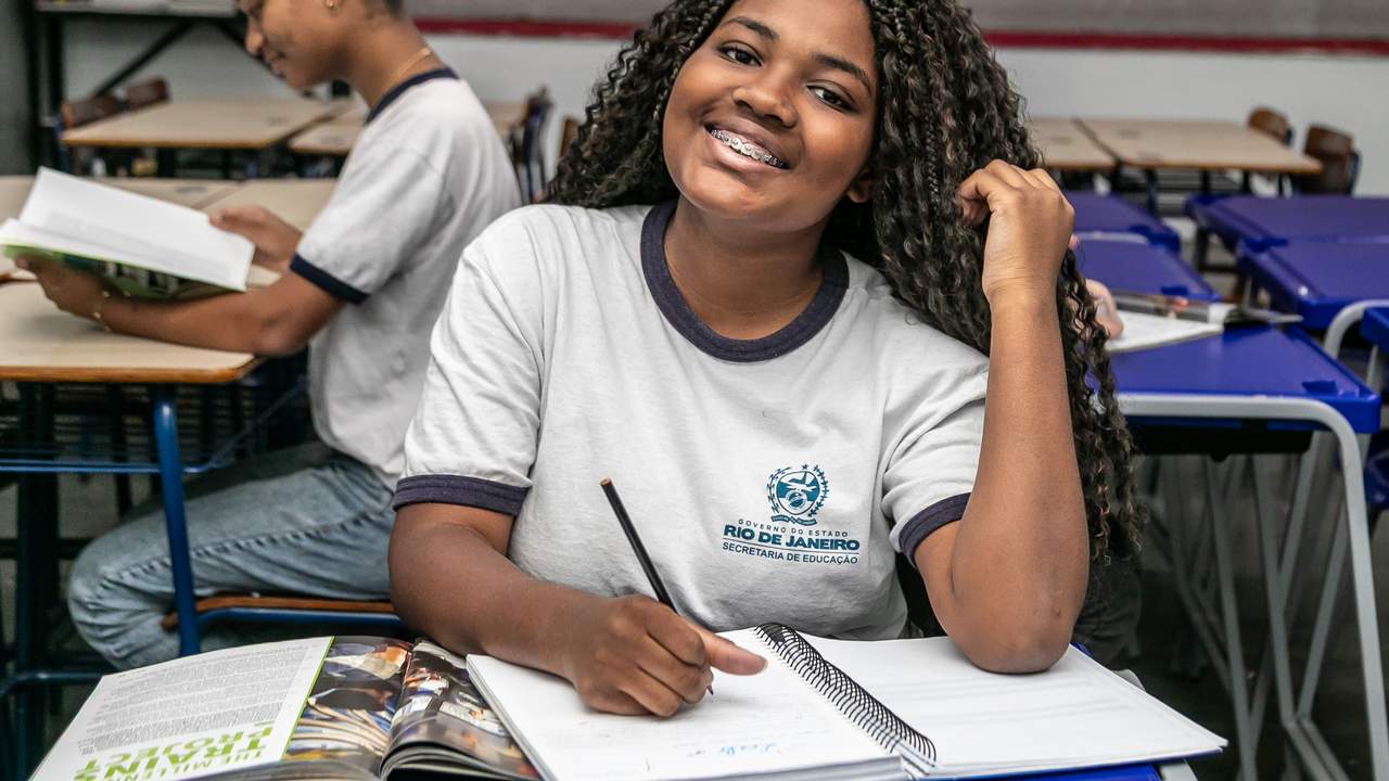 Estudante sorrindo com aparelho nos dentes, em sala de aula, escrevendo em caderno.