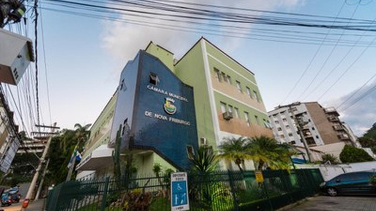 Fachada da Câmara Municipal de Nova Friburgo, Brasil, com céu azul e vegetação ao redor.