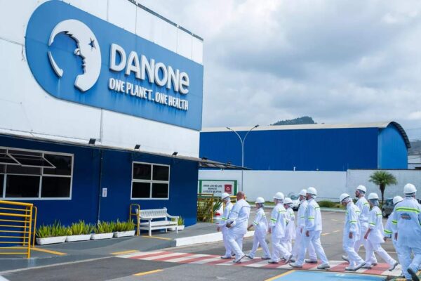 Fachada da fábrica da Danone com funcionários em uniforme branco e o logo da empresa.