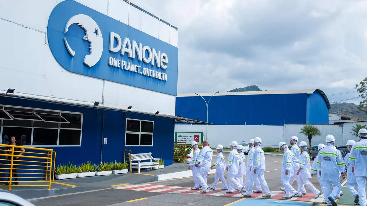 Fachada da fábrica da Danone com funcionários em uniforme branco e o logo da empresa.