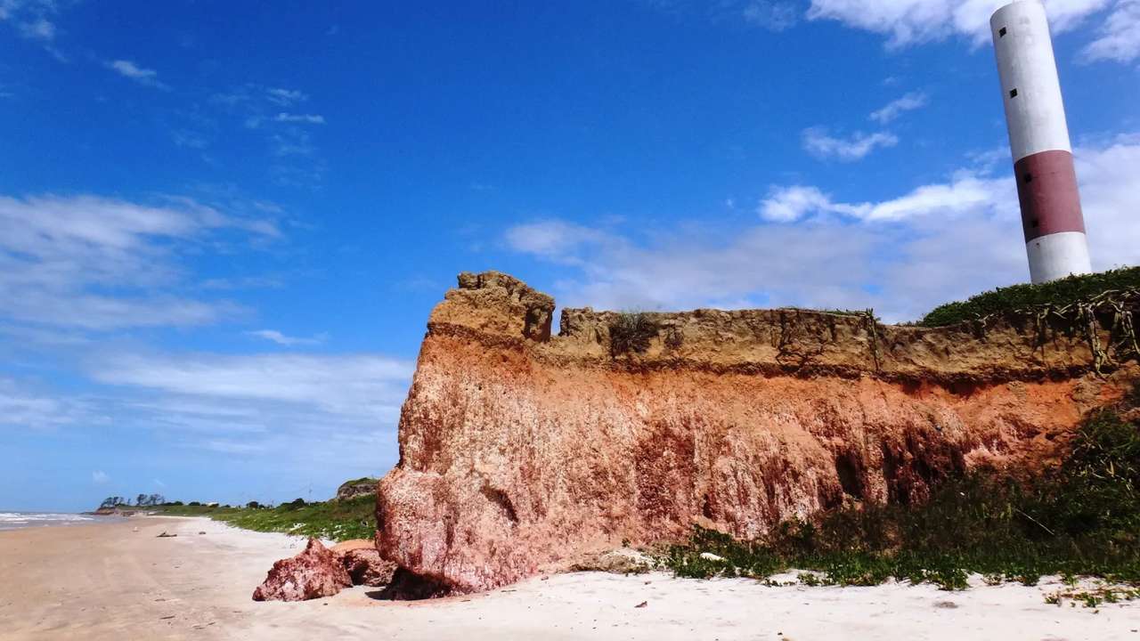 Falésia colorida em São Francisco de Itabapoana, com farol ao fundo e praia.