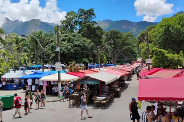 Feira movimentada em Teresópolis com turistas e barracas coloridas, montanhas ao fundo.