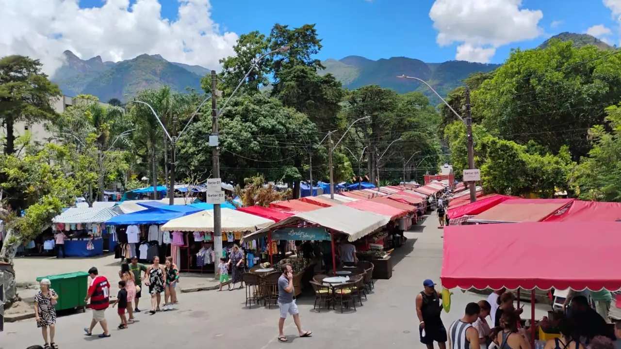 Feira movimentada em Teresópolis com turistas e barracas coloridas, montanhas ao fundo.
