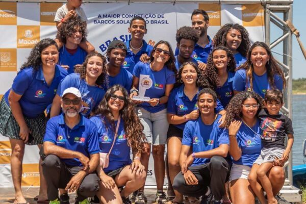 Grupo de estudantes com camisa azul, sorrindo e segurando troféu de competição de barco solar.