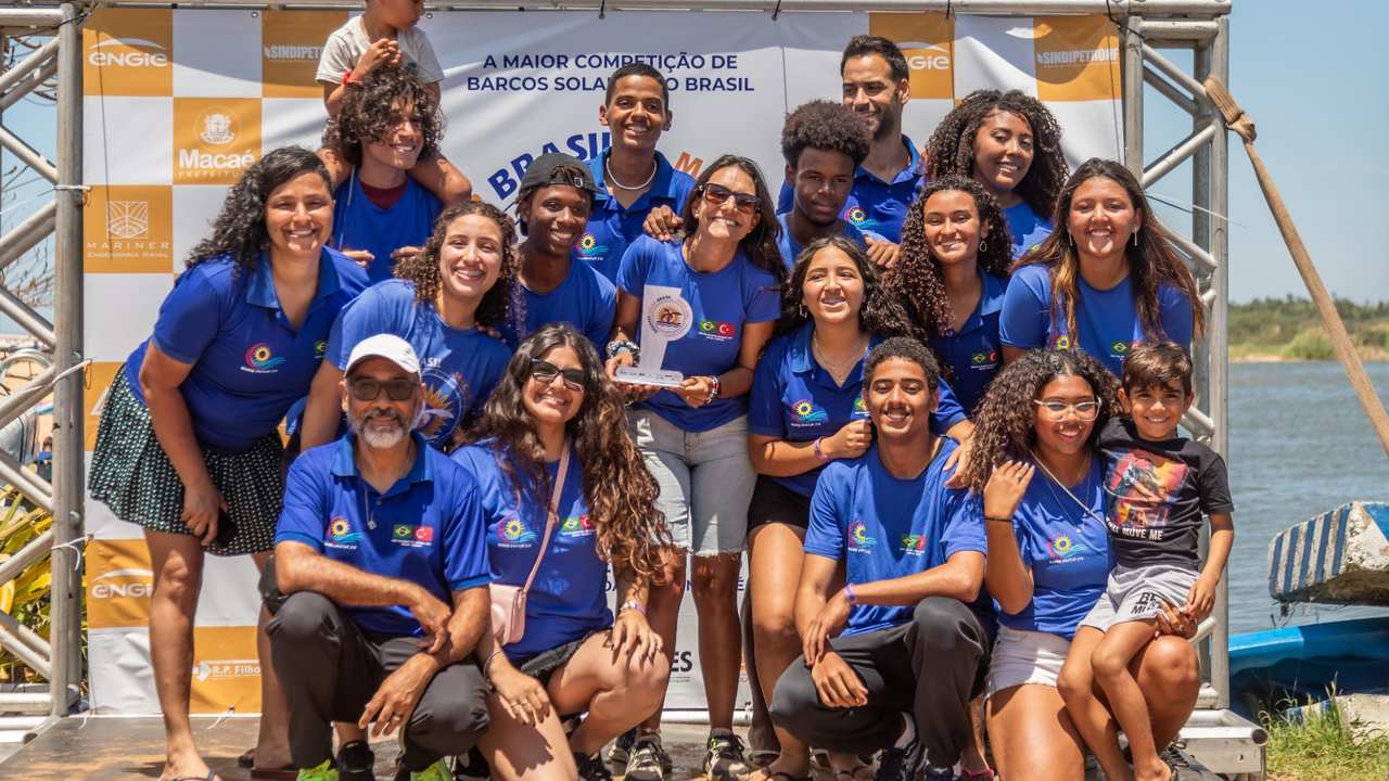 Grupo de estudantes com camisa azul, sorrindo e segurando troféu de competição de barco solar.