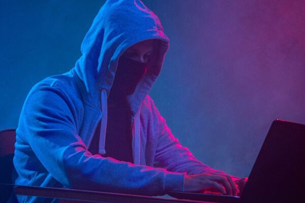 Hacker com capuz e máscara operando um laptop sob luzes de neon.