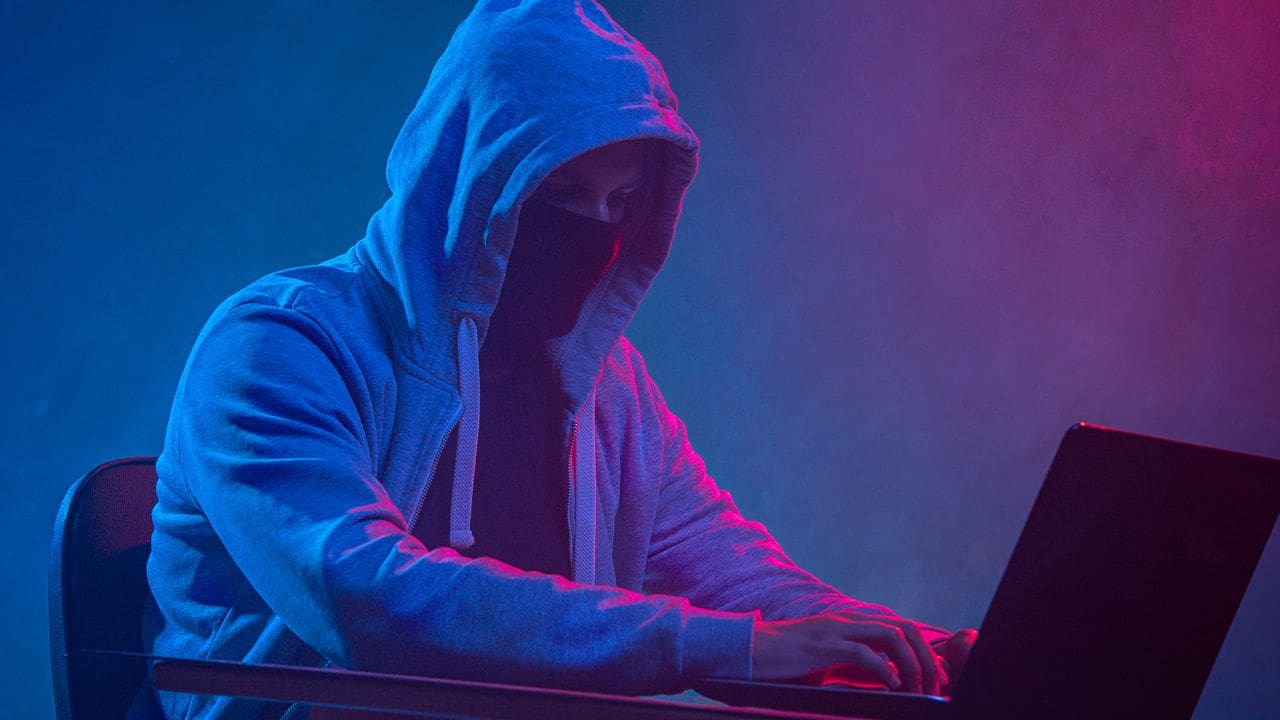 Hacker com capuz e máscara operando um laptop sob luzes de neon.