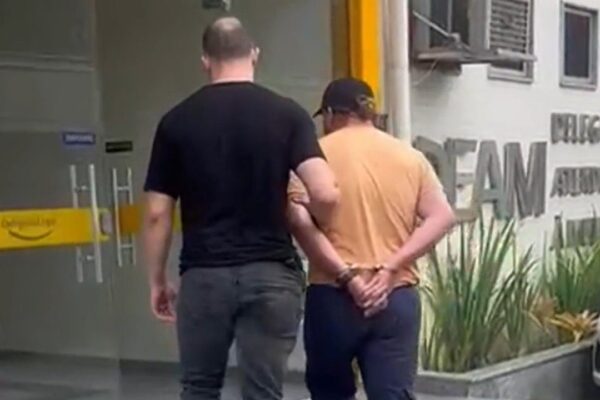 Homem algemado sendo escoltado por policial em frente à delegacia DEAM-NF.